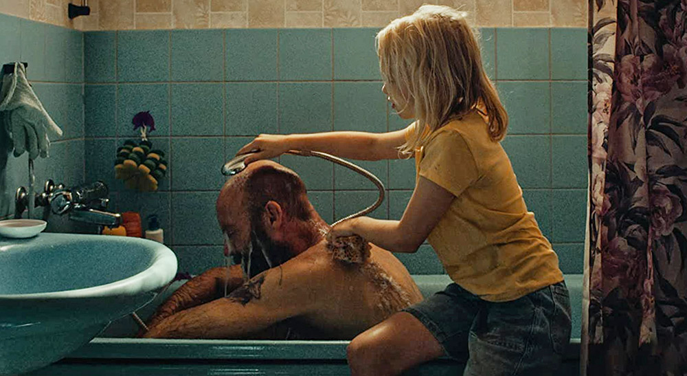En man sitter i ett badkar och blir duschad av en liten tjej.