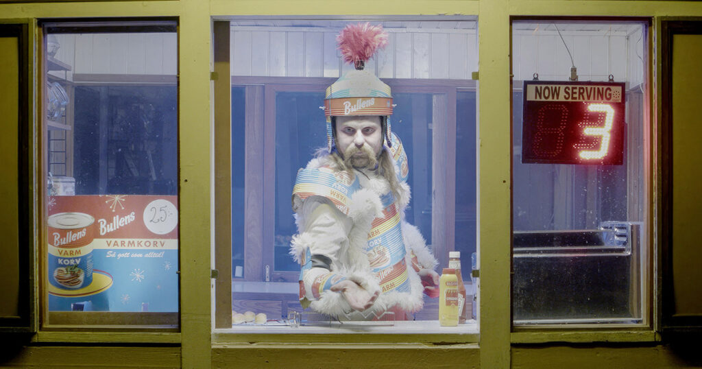 En kille i en korvkiosk, han bär en rustning av Bullens pilsnerkorvburkar.